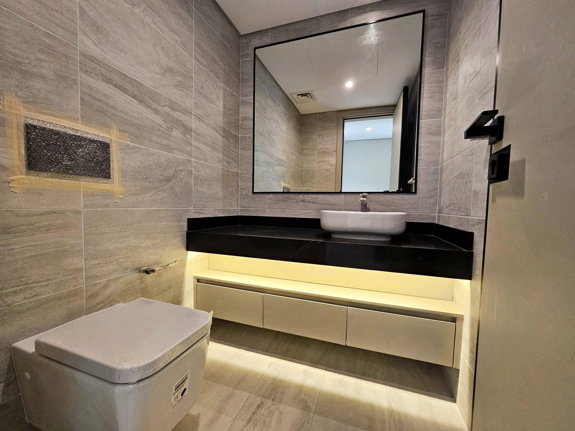 Modern bathroom with gray tiles, a backlit mirror above a white basin on an Auto Draft yellow vanity, and a wall-mounted toilet.