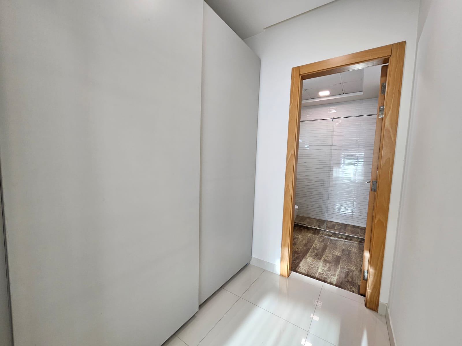 A bright hallway in a luxurious apartment, leading to a room with an open wooden door and frosted glass panels, available for rent in Juffair.