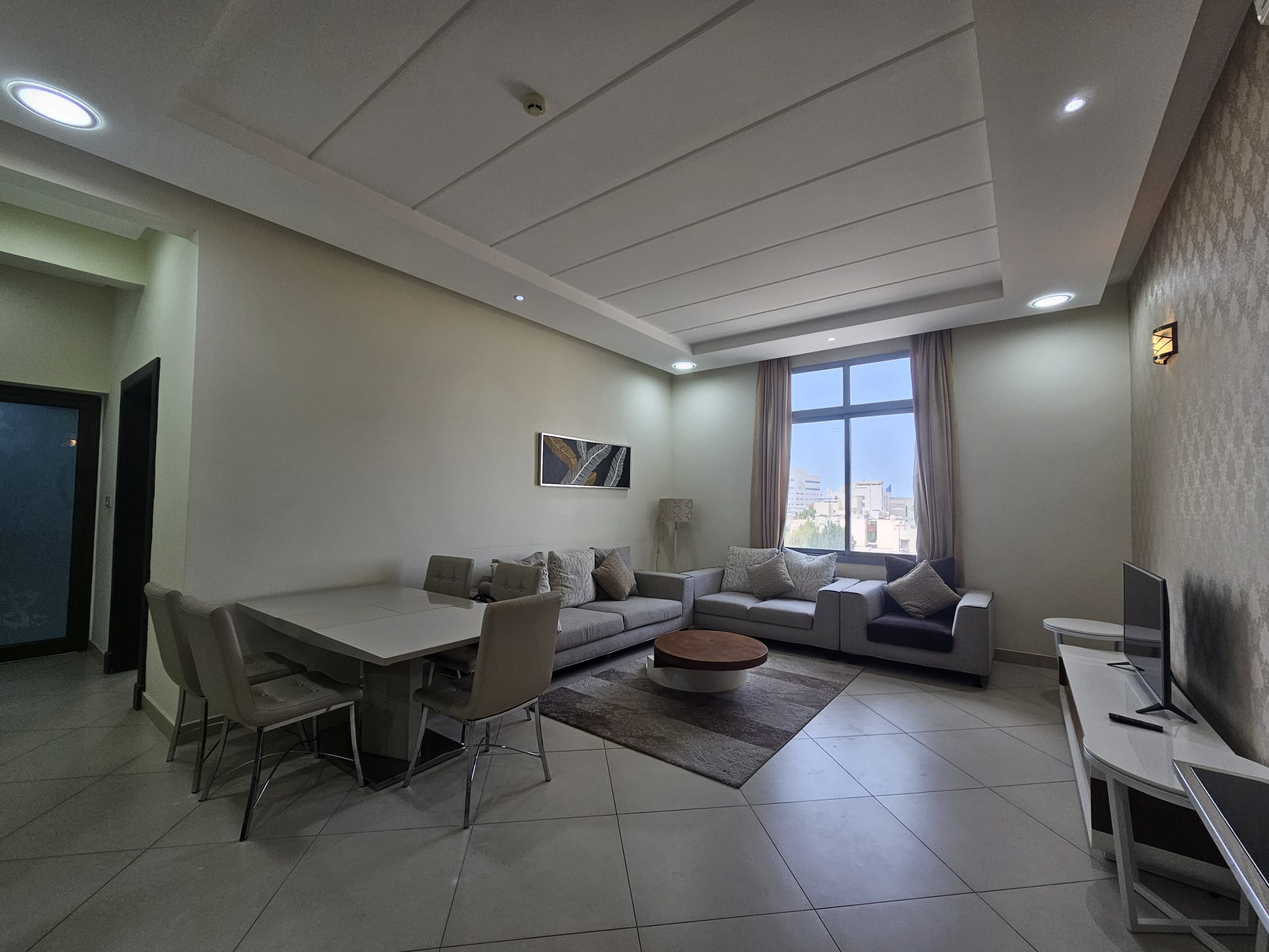 Modern living room with dining area in a 2BR Furnished Flat, featuring a neutral color palette, couch, dining table, and TV, illuminated by natural and artificial light.