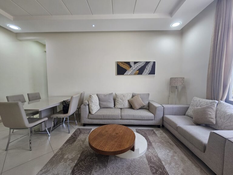 Modern living room in a 2BR Adliya furnished flat with a beige sectional sofa, wooden coffee table, and a dining area in the background.