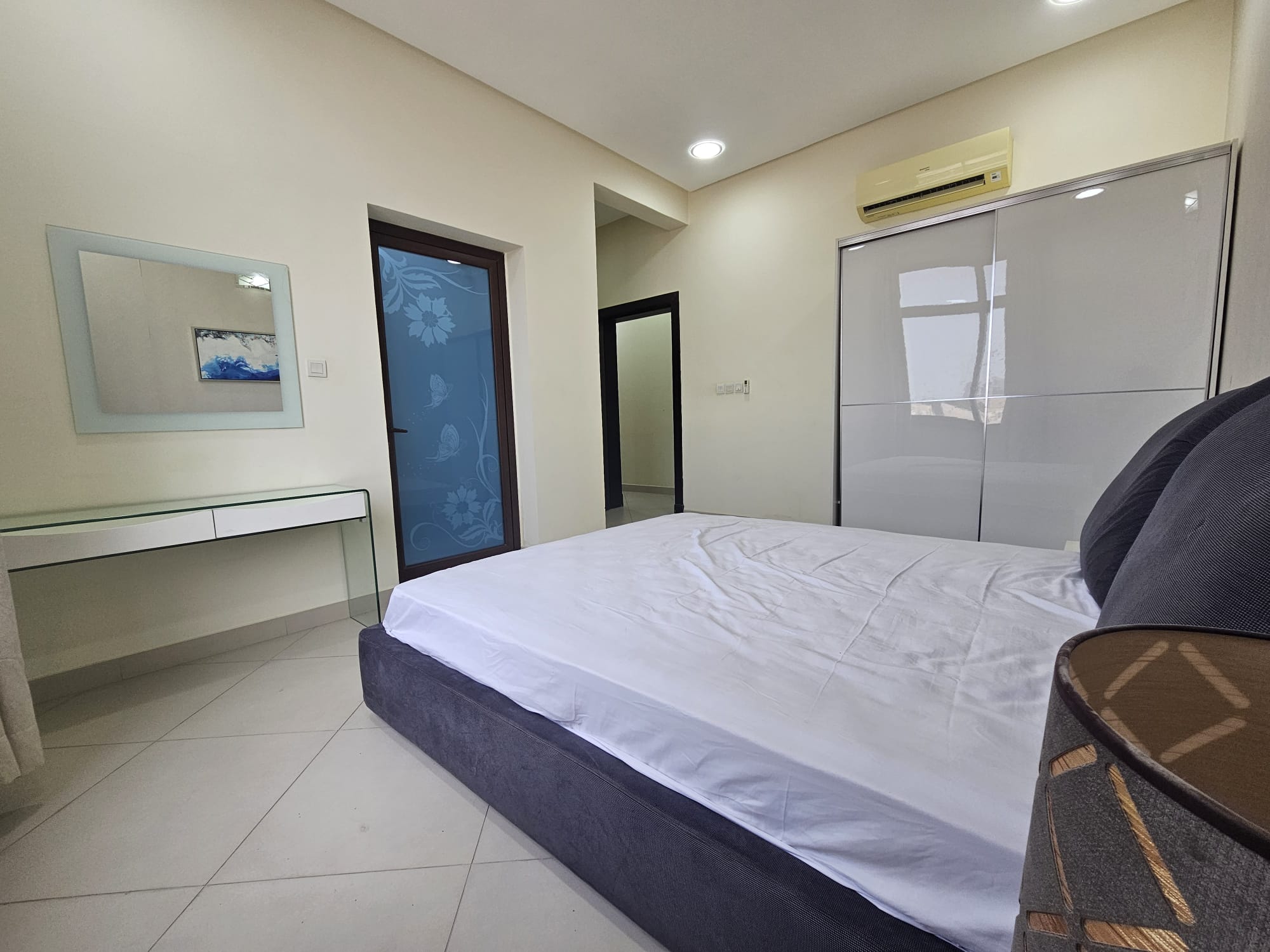 A modern 2BR furnished flat in Adliya with a bedroom featuring a large bed, glass table, air conditioner, and artwork on the wall.