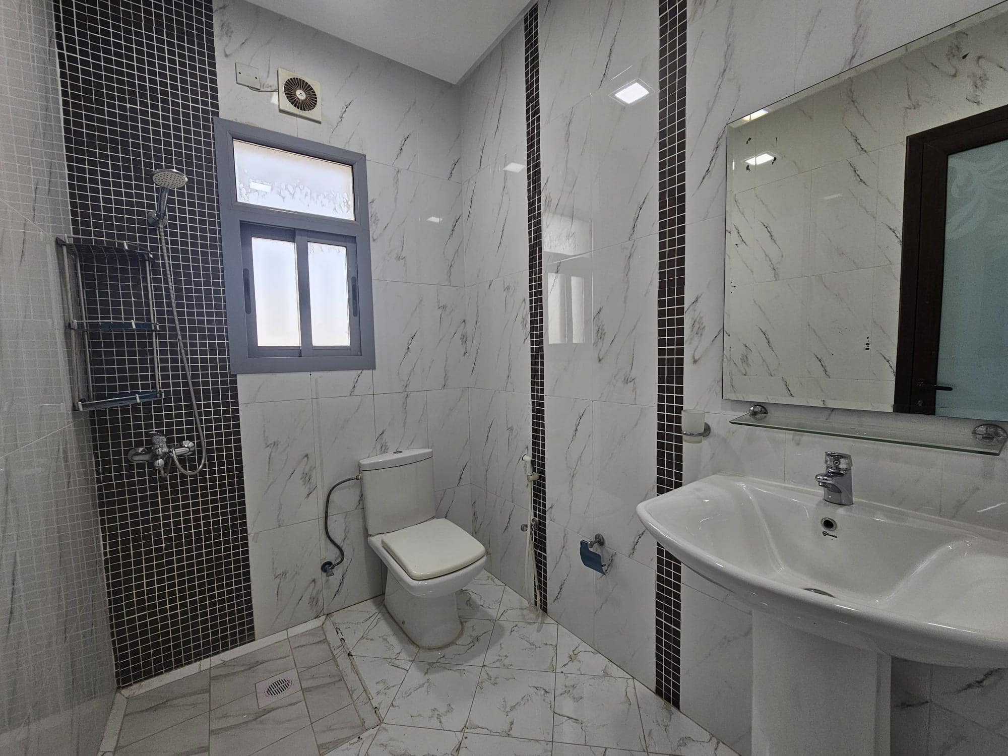 A modern bathroom interior in a 2BR Furnished Flat in Adliya, featuring white and gray marble tiles, a glass shower enclosure, a white toilet, and a pedestal sink.