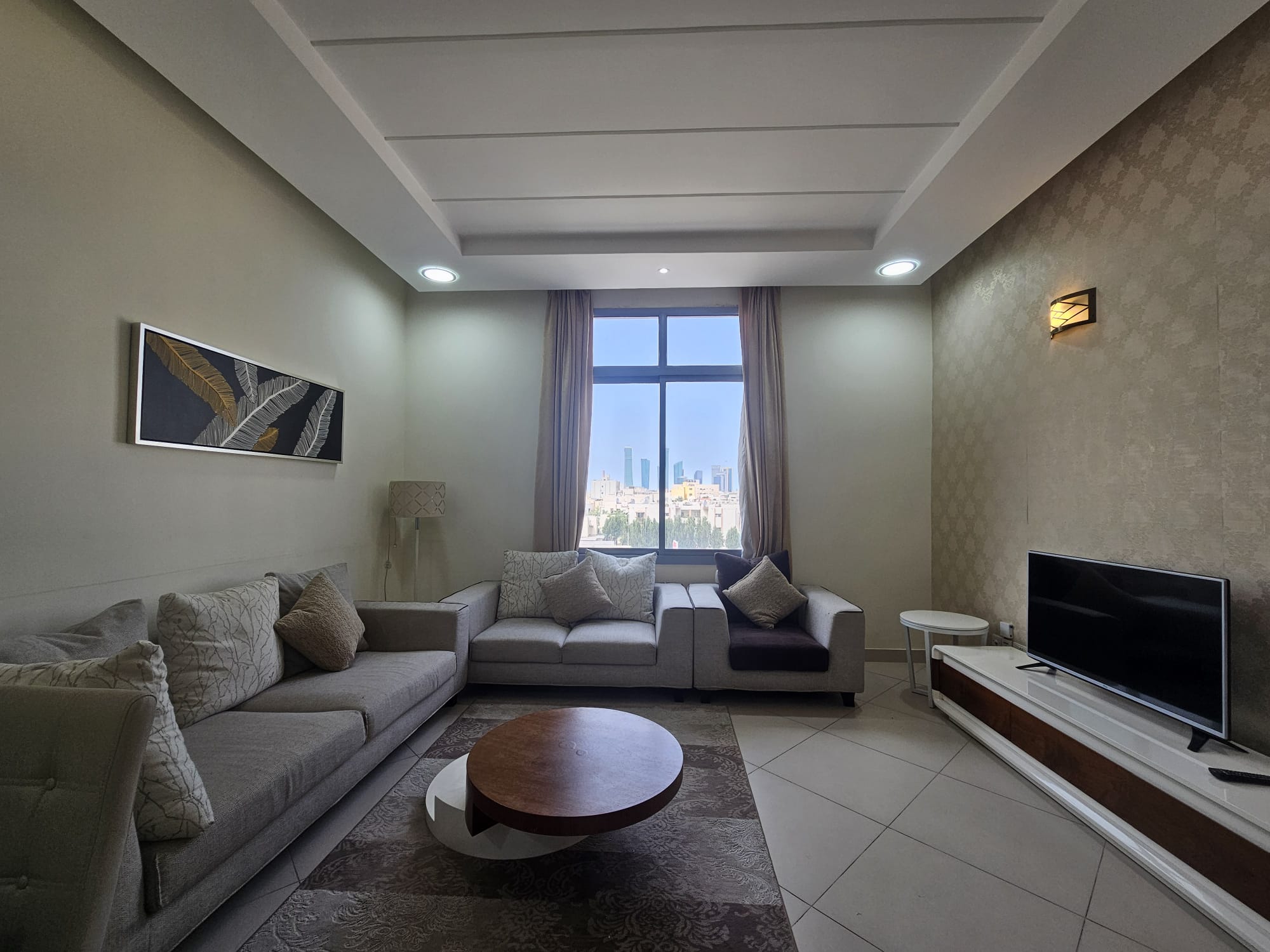 Modern living room in a 2BR Adliya furnished flat with a neutral color scheme, featuring a sofa, armchair, and television, with a cityscape view through the window.