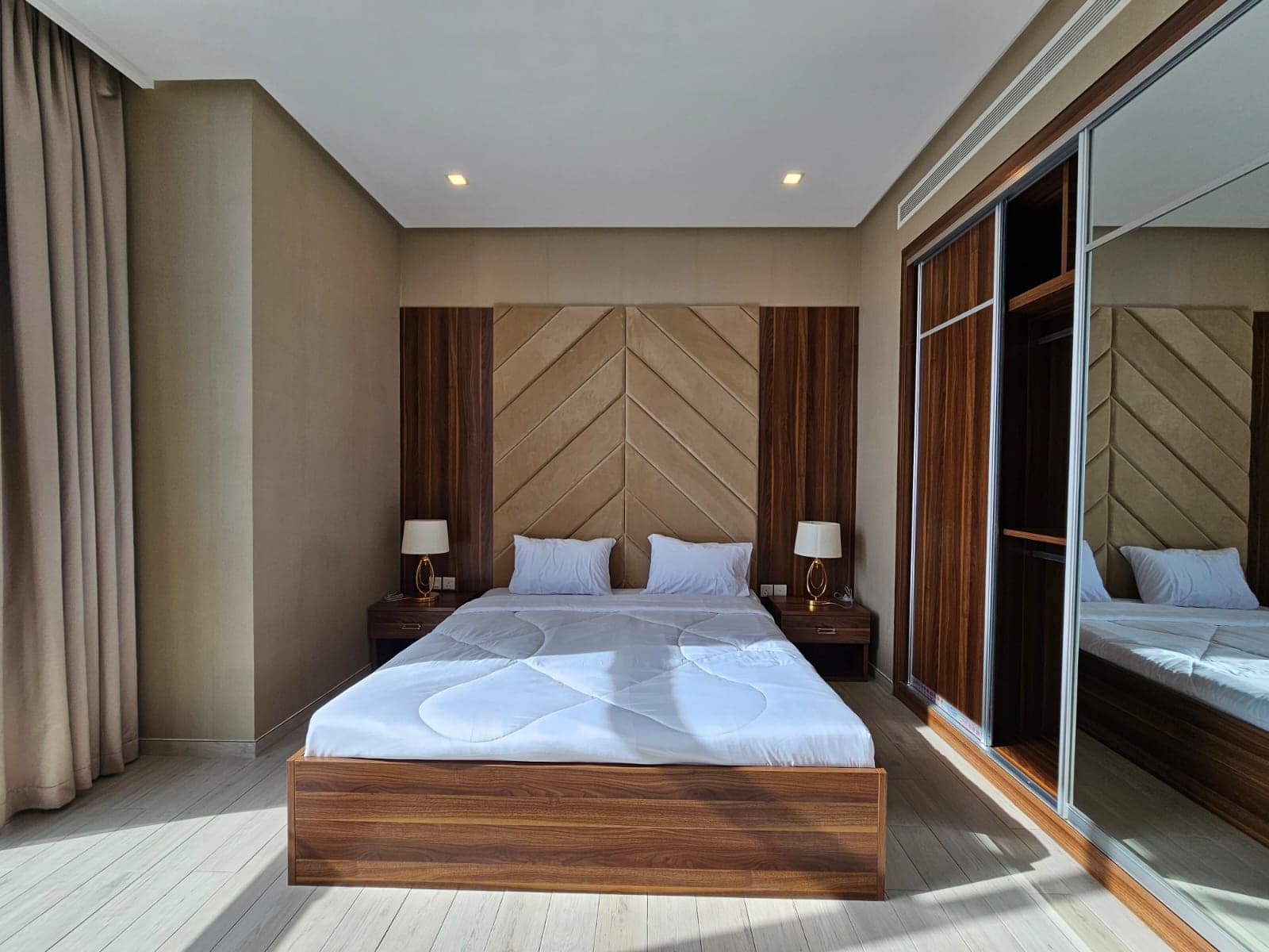 A modern bedroom with a large bed, wooden Auto Draft headboard, bedside tables with lamps, and sliding glass doors leading to an adjacent room.