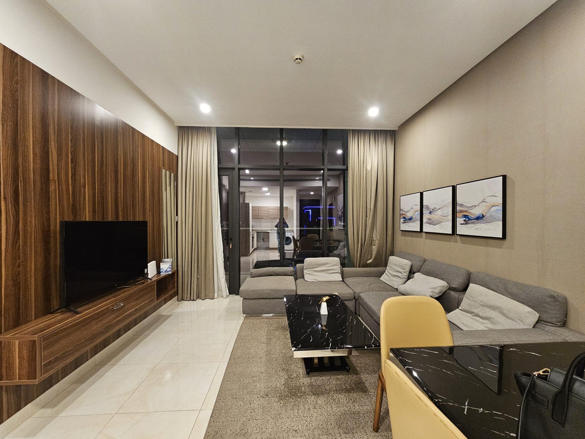 Modern living room with a gray sofa, armchair, black marble coffee table, mounted tv, and wooden panels, leading to a balcony with curtains.