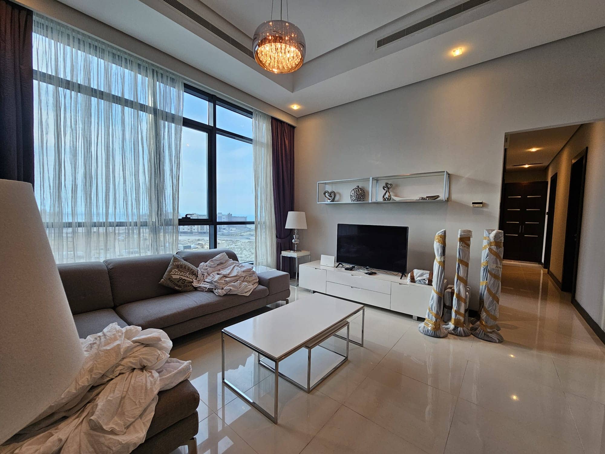 Modern living room interior in a fully-furnished 2 bedroom apartment with ocean view through large window, featuring a beige sofa, white tv cabinet, and glass coffee table. For Rent.