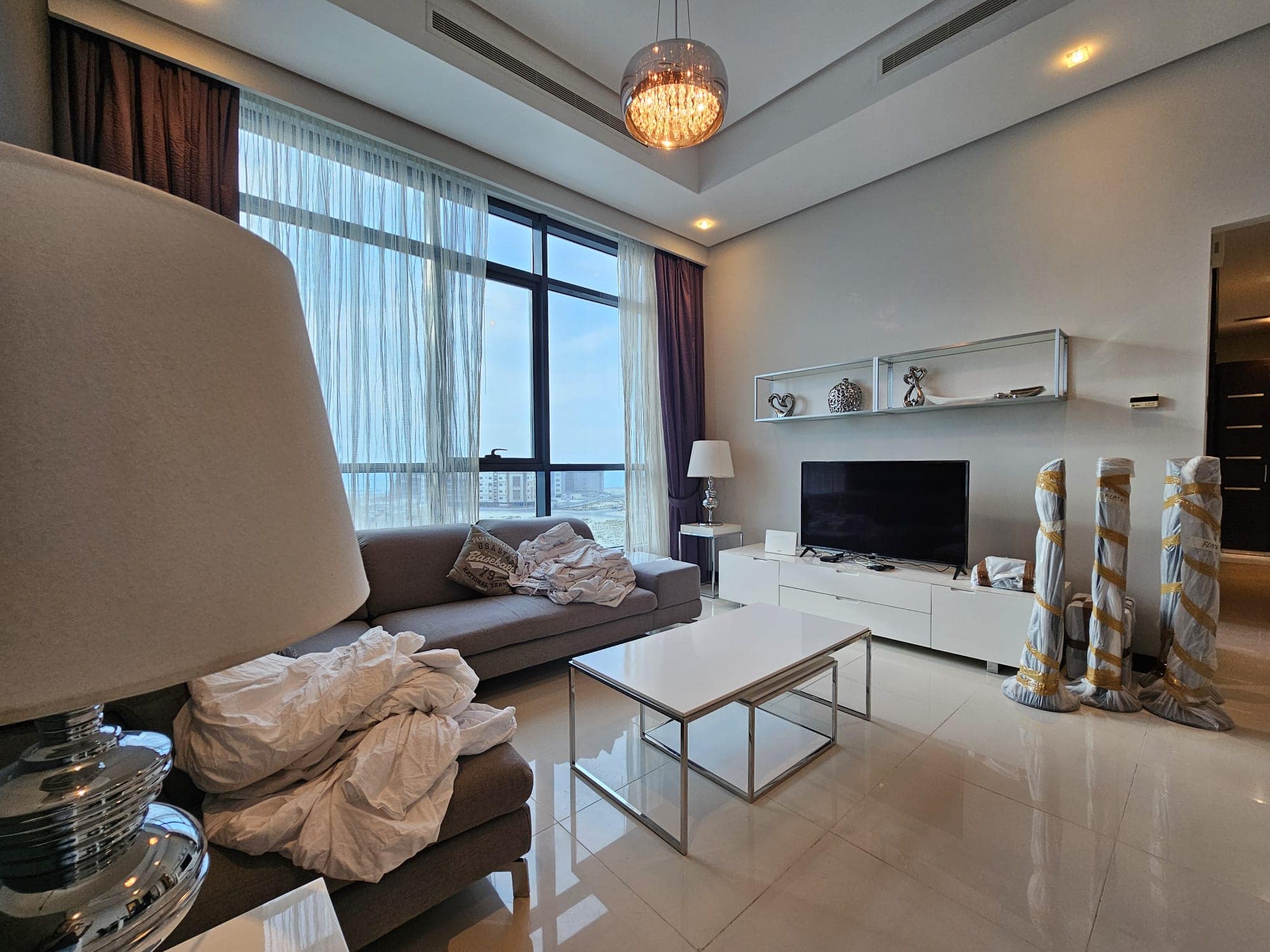 Modern living room with an unmade sofa, glass coffee table, white tv stand, and a large window with ocean view in a fully-furnished 2 bedroom apartment.