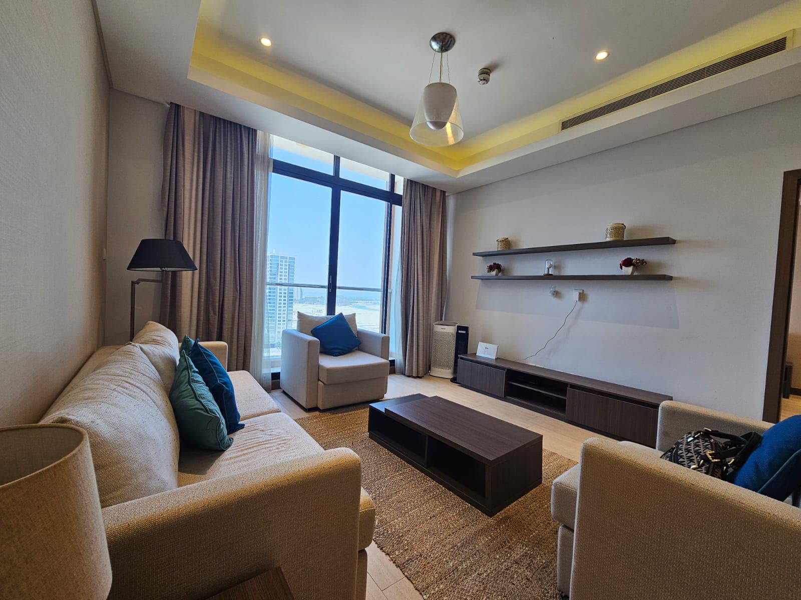 Modern living room with a beige sofa, armchair, wooden coffee table, and shelves against a large window with Auto Draft cityscape views.