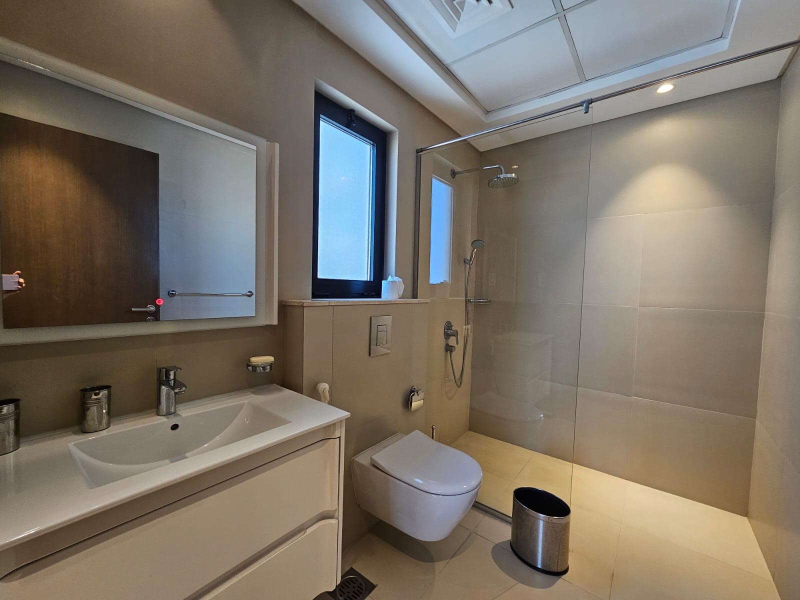 Modern bathroom interior with beige tiles, featuring a sink, toilet, shower area with glass door, and a small window.