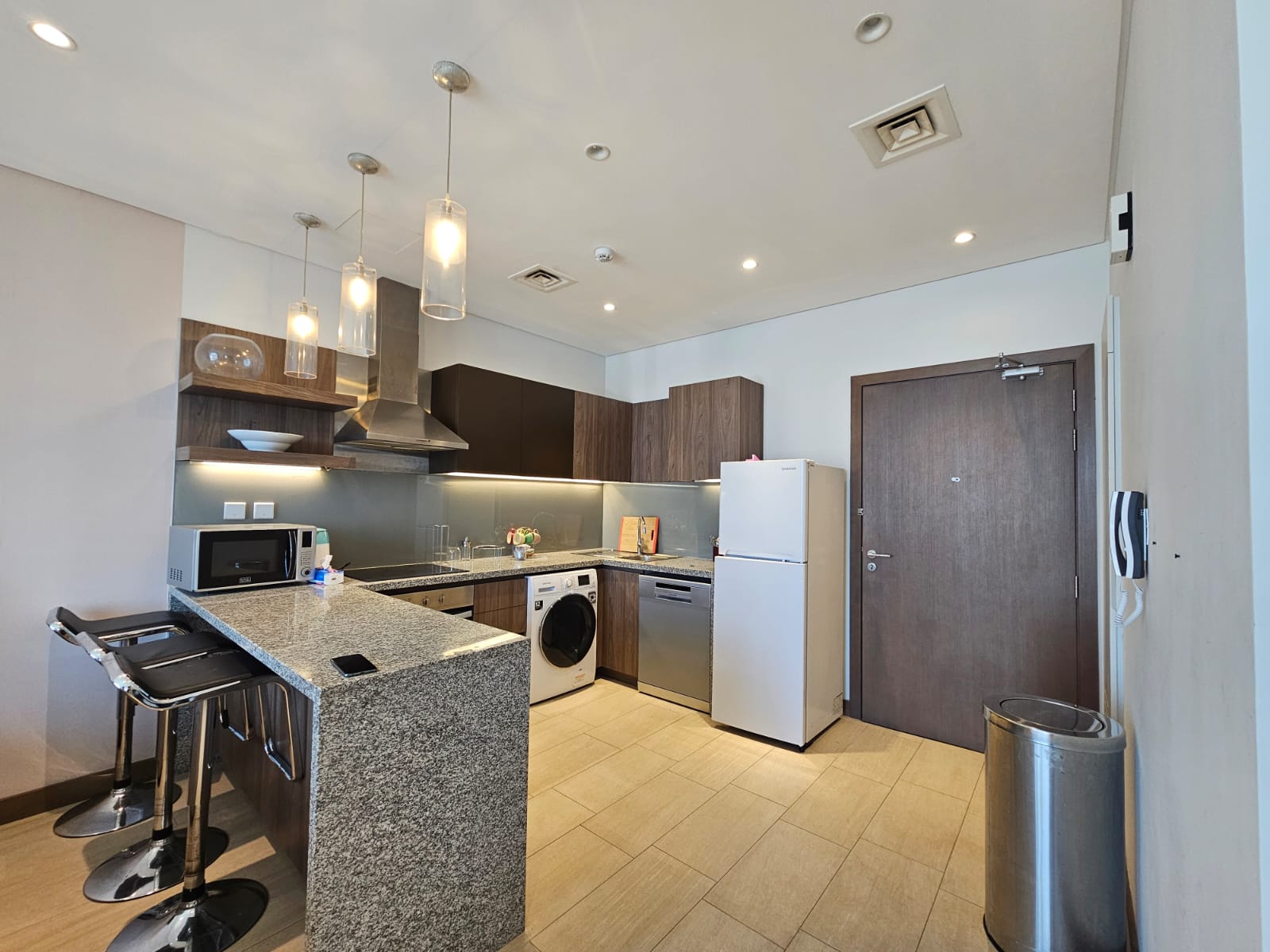 Modern kitchen interior in a Seef flat for rent featuring dark wood cabinets, stainless steel appliances, a white refrigerator, and pendant lights, with a central island.