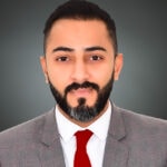 A professional headshot of Maitham Salman with a trimmed beard, wearing a gray suit, white shirt, and red tie, posing against a gray background.
