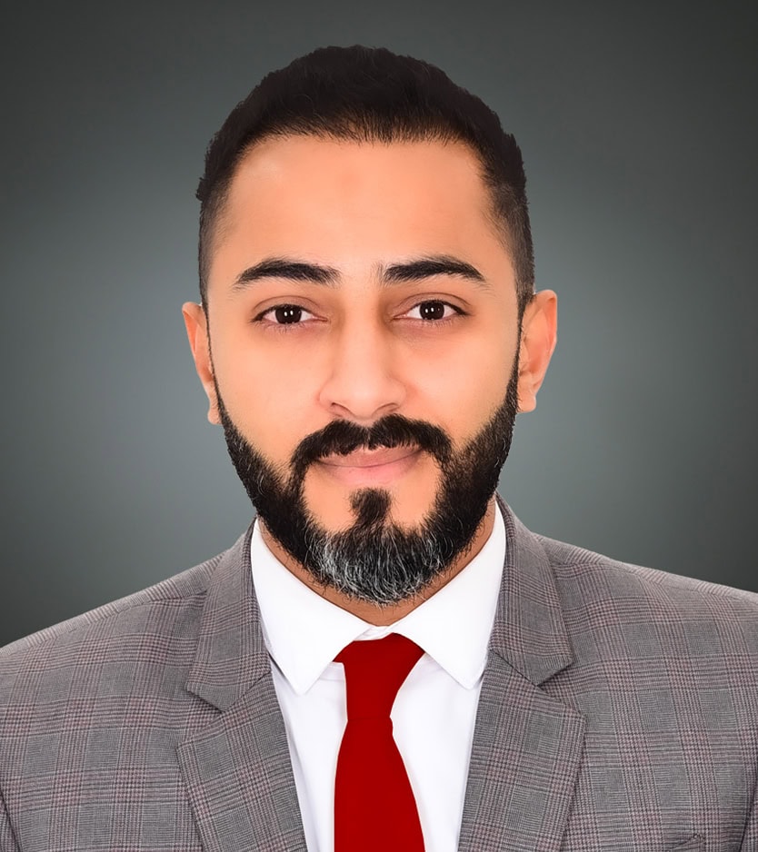 A professional headshot of Maitham Salman with a trimmed beard, wearing a gray suit, white shirt, and red tie, posing against a gray background.