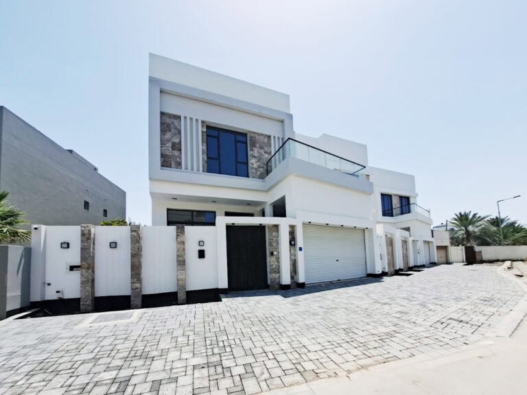 Modern white two-story villa for sale in Saar with a flat roof, large windows, and a cobblestone driveway, flanked by high white walls and garage doors.