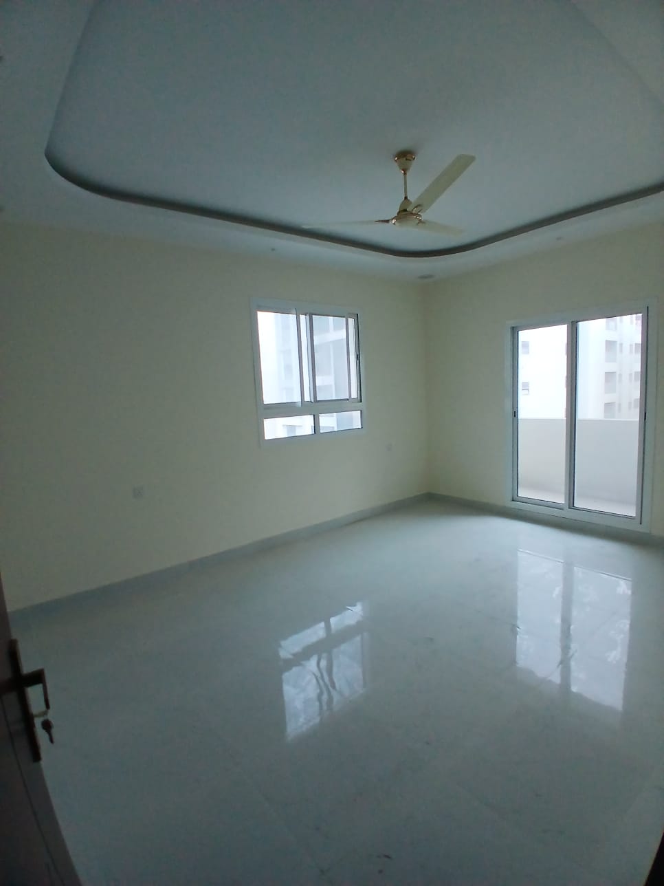 Empty, bright apartment room in a hidden area with white walls, shiny floor, and a corner window next to a ceiling fan.