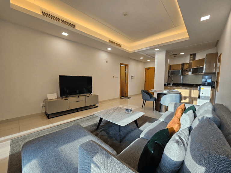 Luxurious apartment living room with an open kitchen layout, available for rent in Juffair, featuring a sofa, coffee table, and television.