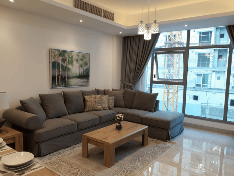 Modern apartment living room with a large gray sectional sofa, wooden coffee table, and a view of the buildings outside in Juffair.