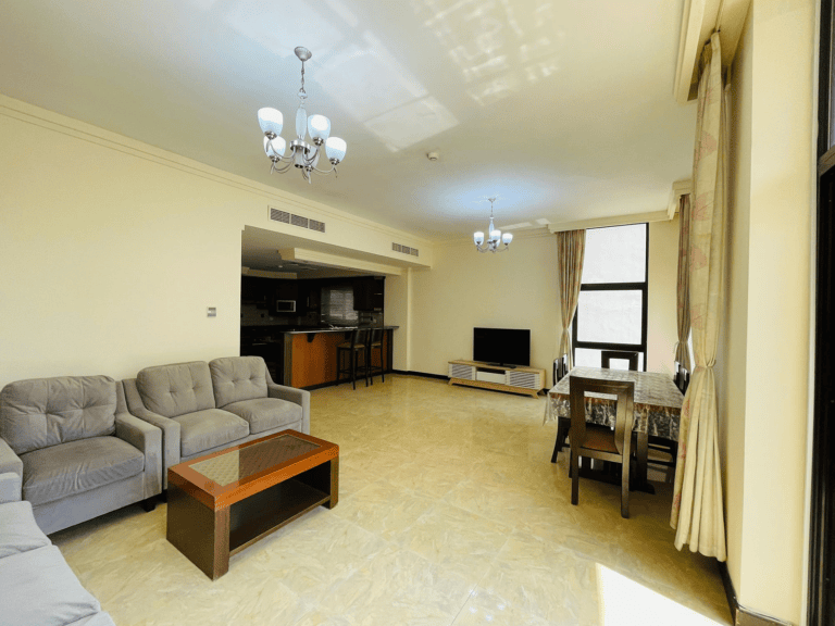 An amazing apartment for rent featuring a brightly lit, spacious living room with a grey sofa set, wooden coffee table, dining area, and an open kitchen in the background, located in Juffair.