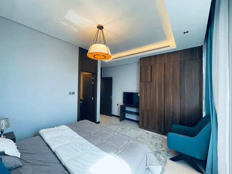 Modern bedroom interior in an amazing apartment for rent in Juffair, featuring a large bed, wood-finished furniture, and a hanging light fixture with blue accents.