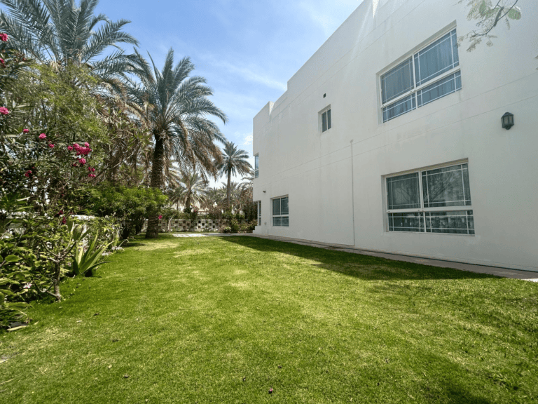 Well-manicured lawn beside a white building with lush palm trees and shrubbery under a clear sky.