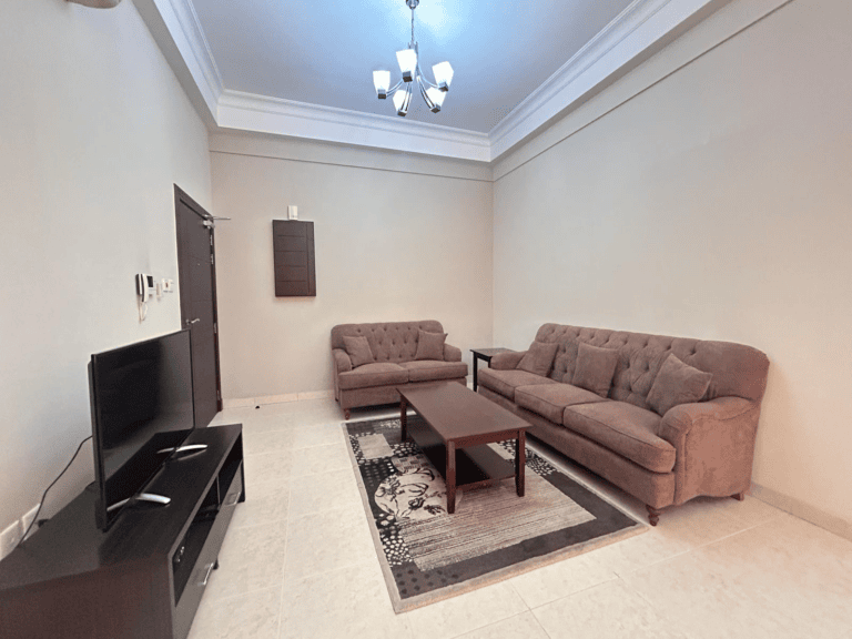 A neatly arranged living room within a luxury 2 BDR Apartment in Saar, with a plush sofa set, a television on a stand, and a coffee table on an area rug, all fully