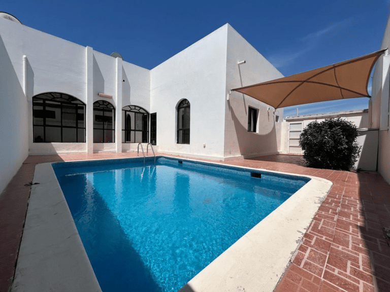 A sunlit outdoor pool area with clear blue water, surrounded by a white building with arched doorways and a tiled patio.