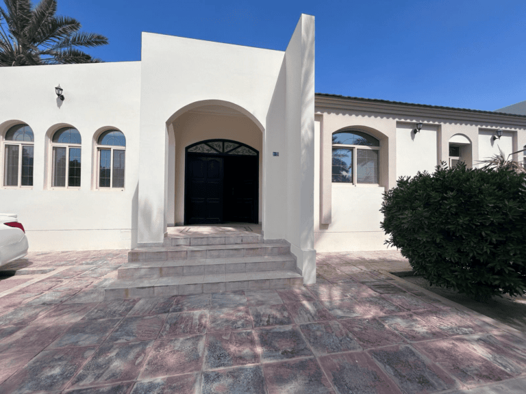 White arched entrance of a semi-furnished villa with a black door, flanked by palm trees and a red-tiled courtyard.