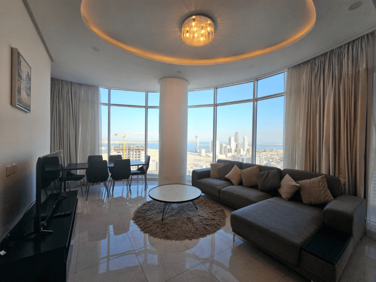 Modern living room in a high-rise apartment in Juffair with a city view, featuring a sectional sofa, circular table, dining area, and ambient lighting.
