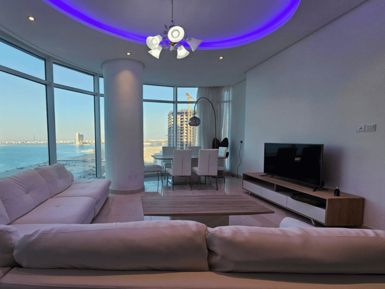 Modern living room with large windows offering a sea view, white sofas, a dining area, and ambient purple ceiling lighting.