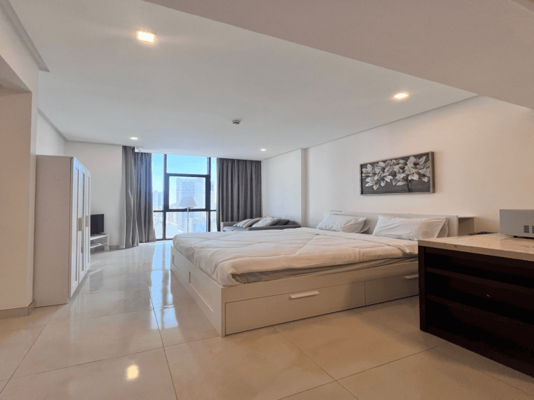 Modern flat for sale with a large bed, glossy tile floor, white walls, and a city view through a window in Juffair.