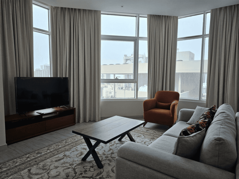 Modern flat for rent with a living room featuring a gray sofa, an orange armchair, a black TV on a wooden stand, and large windows with city views.