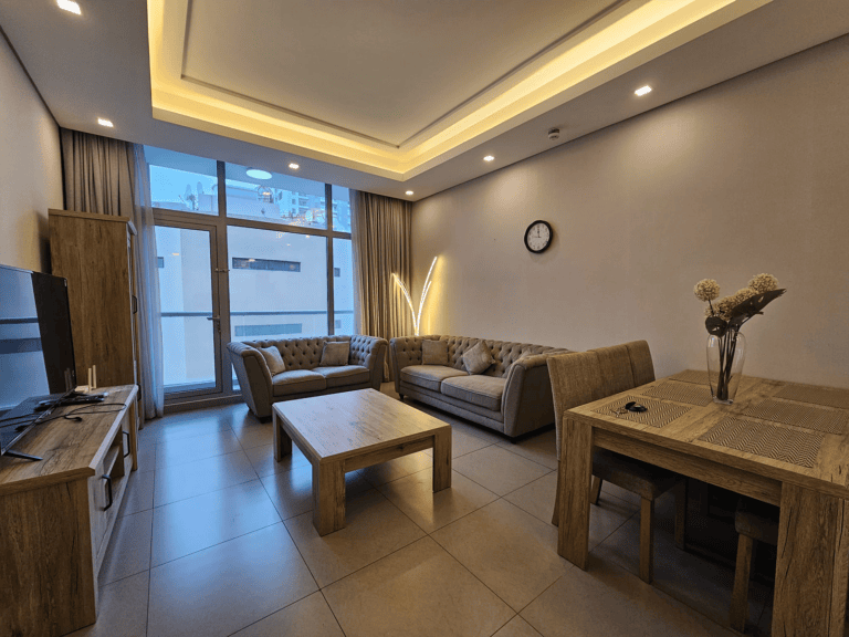 Modern living room with sectional sofa, wooden coffee table, LED lighting, and large windows with blinds in a fully furnished 2-bedroom apartment in Juffair.