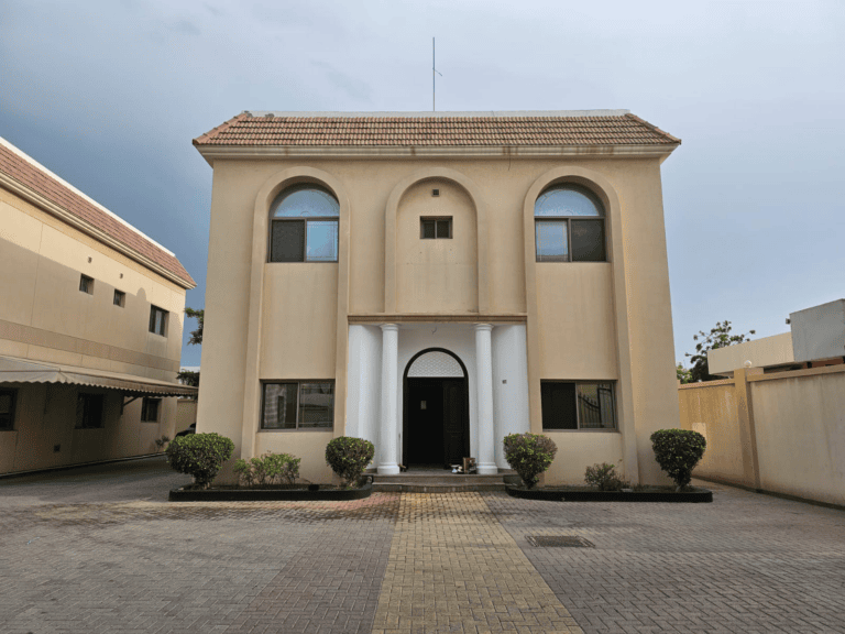 Two-story beige villa for rent in Juffair with arched doorway, symmetrical windows, and tiled pathway, flanked by small shrubs under a cloudy sky.