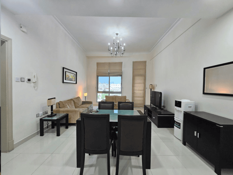 Modern 2-bedroom apartment in Adliya with a living room featuring a dining set, sofa, and TV, illuminated by natural light and a chandelier, with a neutral color scheme.