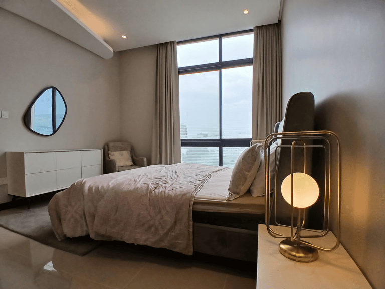 Luxury bedroom with a bed covered in a gray quilt, a round mirror on the wall, and a stylish floor lamp beside a large window with curtains.