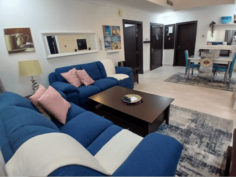Bright modern living room with a blue and white sofa, a large coffee table, and a dining area in the background in a flat for rent in the Busaiteen area.
