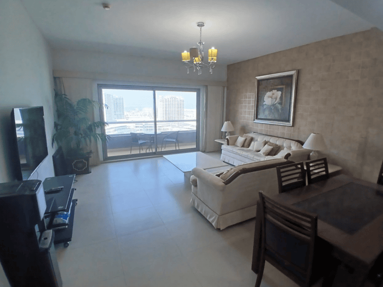 Modern living room in an Amwaj flat with a cream sofa, dining area, and a large window with a city view, accented by tiled walls and sleek furnishings.