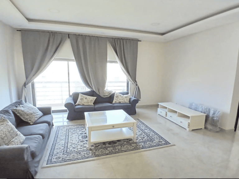 A bright modern living room in a three-bedroom apartment with a gray sofa, patterned cushions, a white coffee table, an area rug, and long curtains in front of a large window.