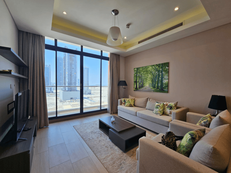 Modern living room in a Seef flat, furnished with a beige sofa, patterned cushions, and a flat screen TV. Large windows offer views of city buildings.