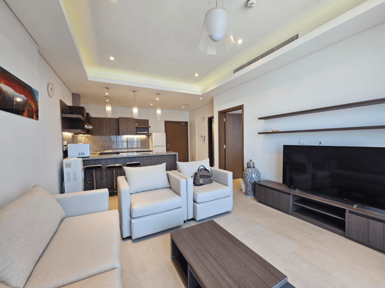 Modern flat for rent featuring a living room with white sofas, a large TV, and an open-plan kitchen with dark cabinetry and stainless steel appliances.