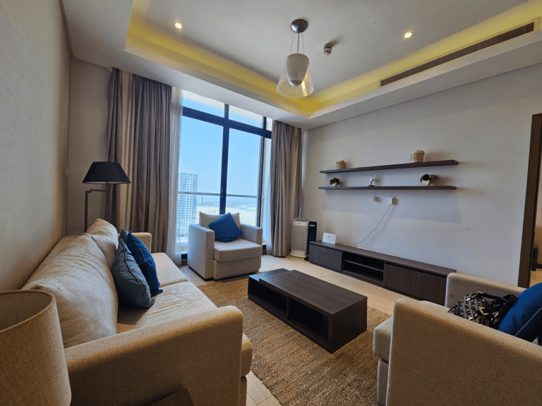 Modern living room with a beige sofa, armchair, white ottoman, wooden tv stand, floating shelves, and large windows overlooking a cityscape in a flat for rent in Seef.