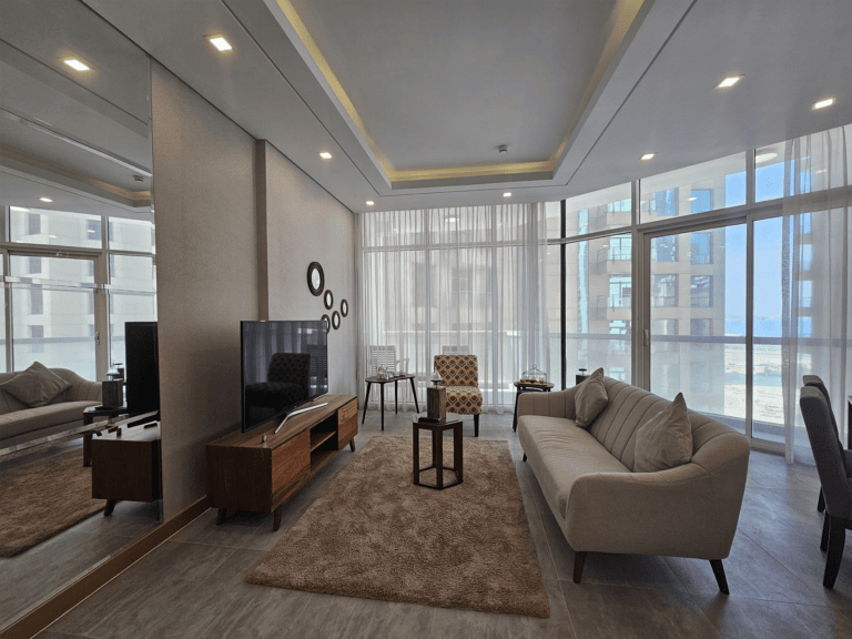 Modern living room in Juffair with neutral tones, featuring a sofa, armchairs, and a TV, with large windows providing ample natural light.