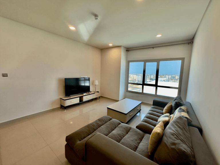Luxury apartment with a modern living room featuring a large sectional sofa, television stand, coffee table, and floor-to-ceiling windows overlooking the cityscape of Janbiyah.