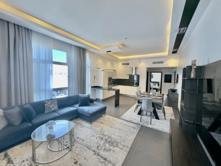 Modern, spacious living room in a luxury apartment with a sectional sofa, glass coffee table, dining area, and kitchen, featuring light color tones and large windows.