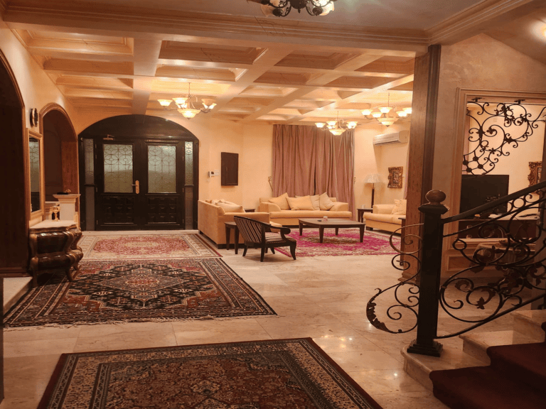 Elegant Villa for Rent in Busaiteen with cream sofas, ornate rugs, wooden ceiling beams, and wrought iron staircase railing.