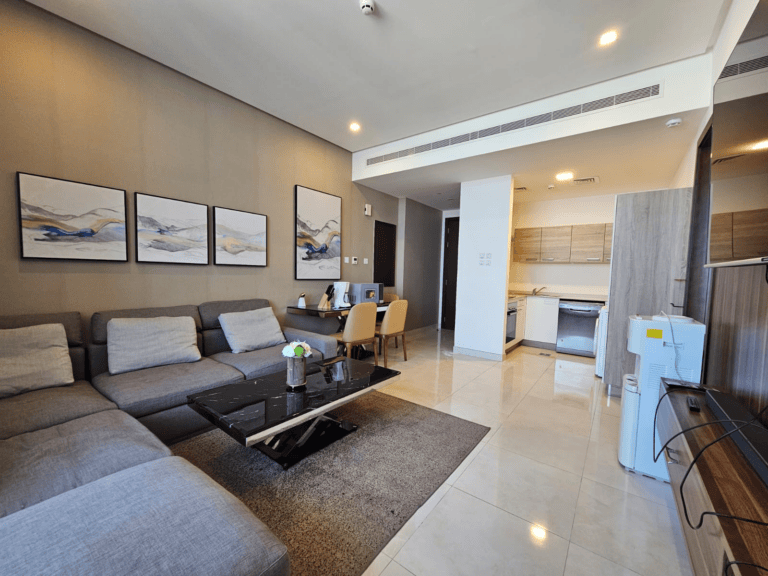 Modern apartment living room with a gray sofa, coffee table, and wall art in Juffair, leading to an open kitchen and dining area.