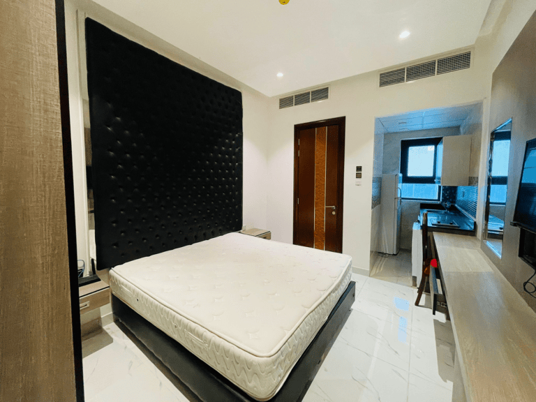 Luxury studio apartment featuring a large black tufted headboard, white mattress on a platform bed, glossy tiled flooring, and an open doorway leading to a bathroom.