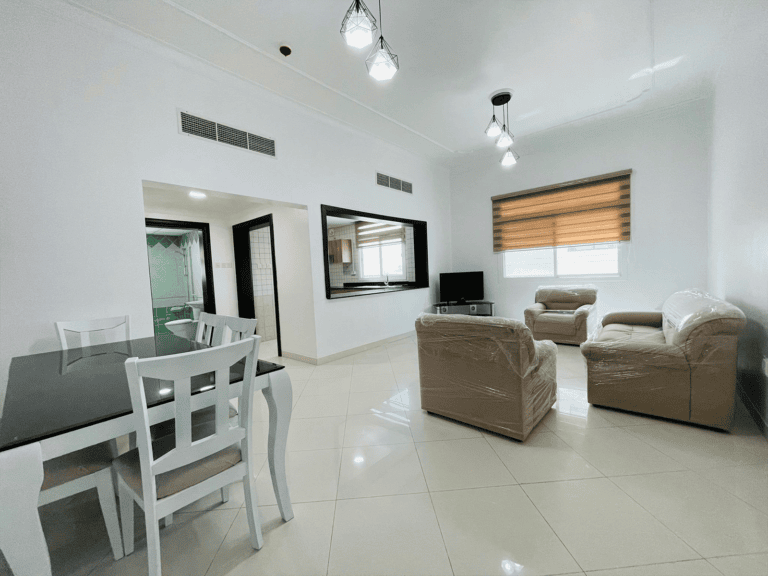 Stylish apartment in the Al-Burhama area with a modern living room featuring a dining area, including a gray table and chairs, cream sofas covered in plastic, and a flat-screen TV