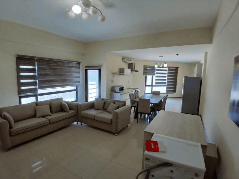 A spacious, modern apartment living room in Al Hidd with beige sofas, a dining area, and an open kitchen, decorated in neutral tones and natural light.