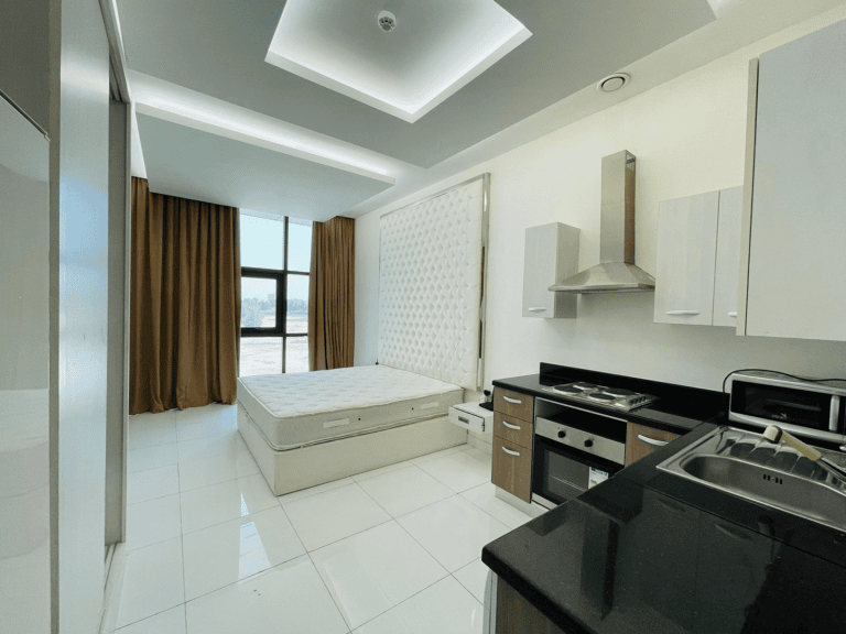 Luxury studio apartment interior featuring a bed, kitchen with black countertops, white cabinets, and large windows with curtains.