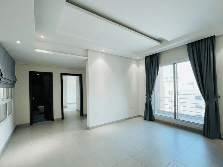 Modern empty living room with white walls, large windows with grey curtains, and tile flooring. Doorways lead to adjacent rooms. Apartment available in Al Burhama.